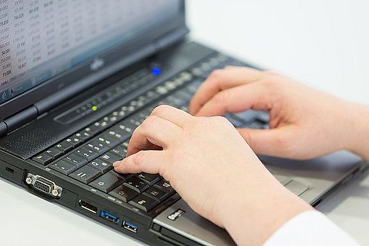Hände an der Tastatur eines Laptops. Auf dem angeschnittenen Bildschirm sind Zahlenkolonnen zu sehen